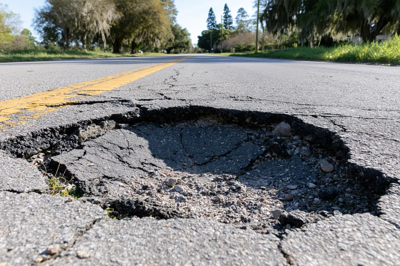 Large pothole with crumbling asphalt edges centered in a two-lane road lined with trees under daylight