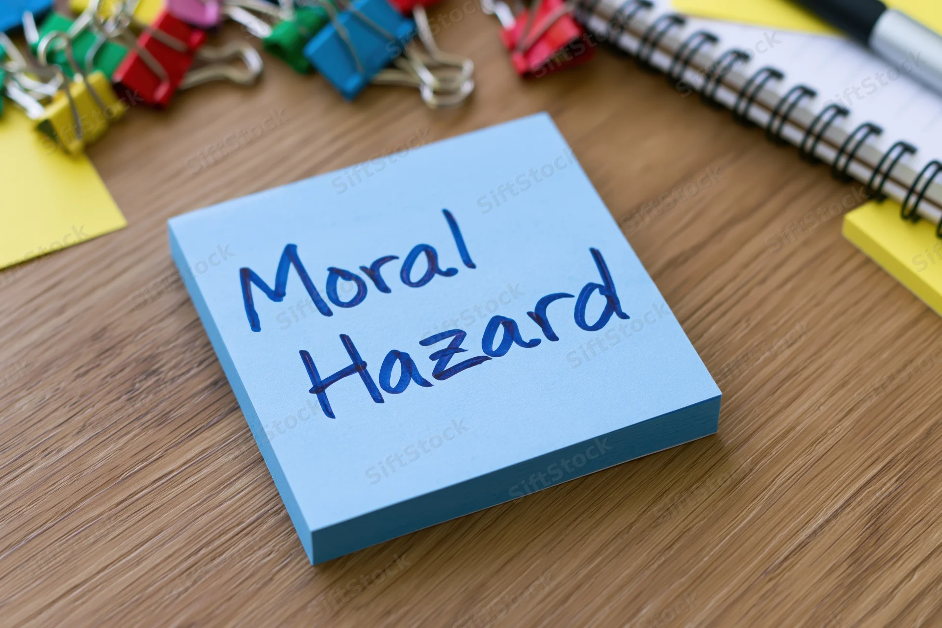 Blue sticky note with handwritten text moral hazard on wooden desk surrounded by office supplies