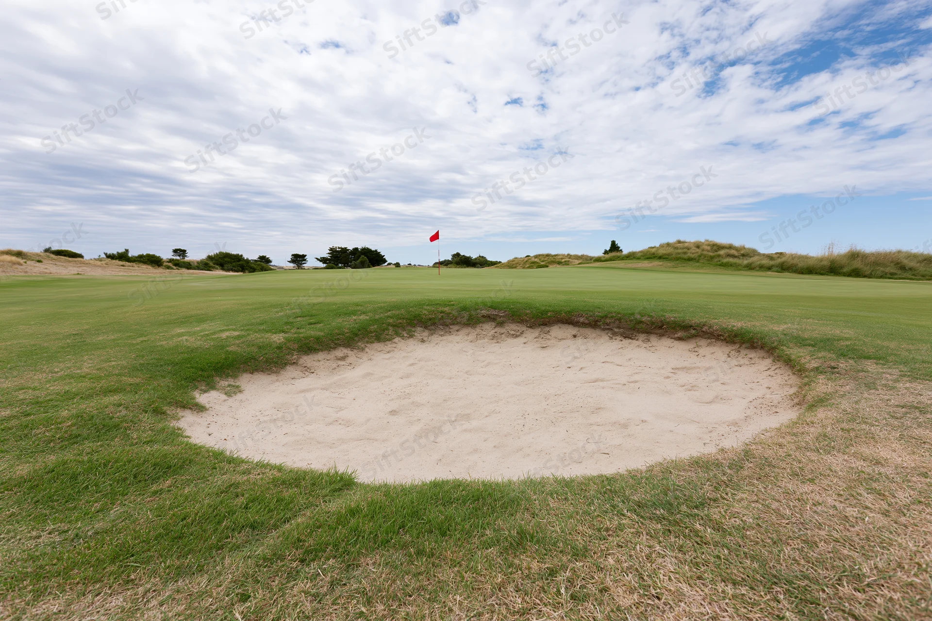 Golf course green with a deep sand bunker in the foreground under a partly cloudy sky