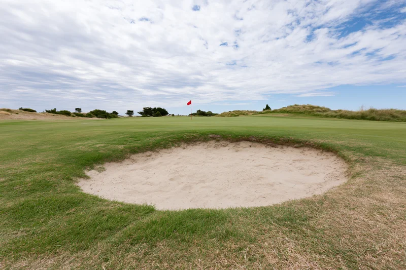 Golf course green with a deep sand bunker in the foreground under a partly cloudy sky