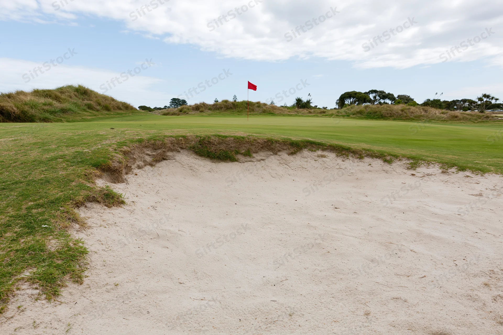 Golf course green with a deep sand bunker in the foreground under a partly cloudy sky