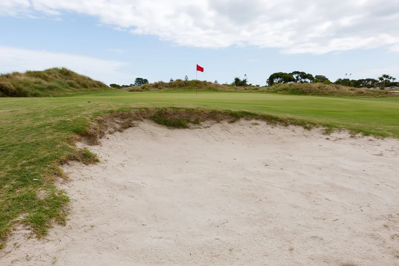 Golf course green with a deep sand bunker in the foreground under a partly cloudy sky