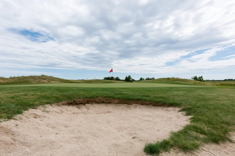 Golf course green with a deep sand bunker in the foreground under a cloudy sky