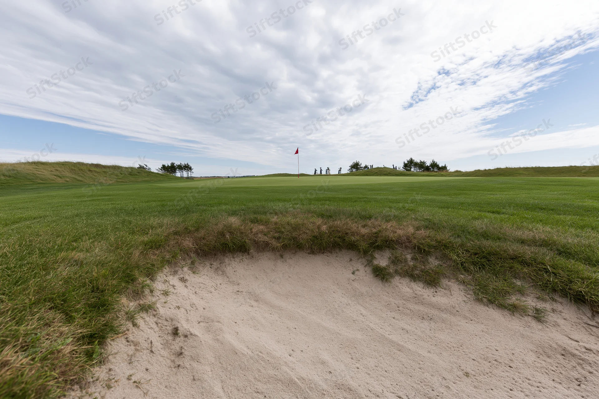 Golf course green with a deep sand bunker in the foreground and players in the distance under a cloudy sky