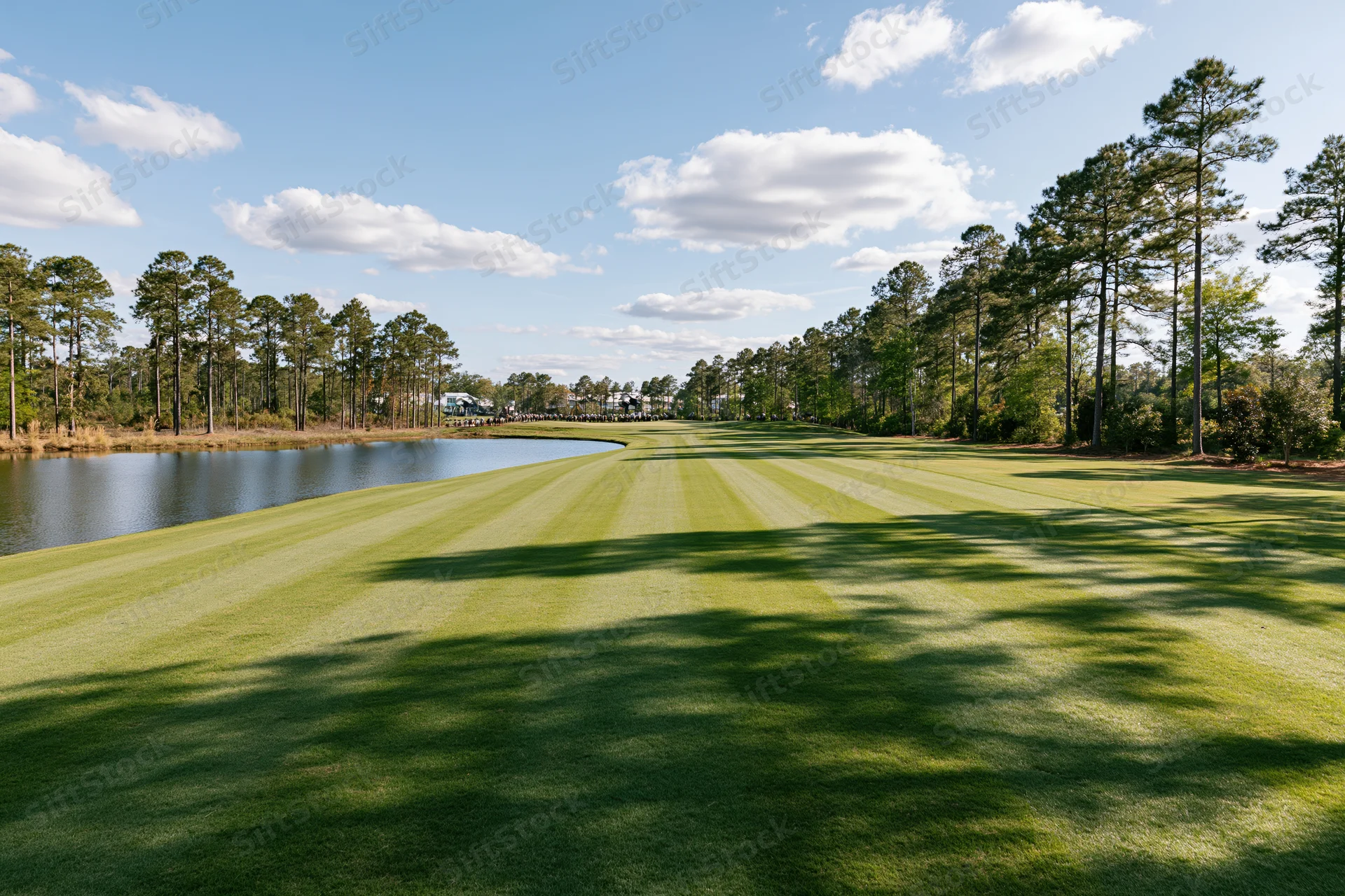 Manicured golf course fairway stretching toward a calm pond lined with pine trees under a blue sky with scattered clouds