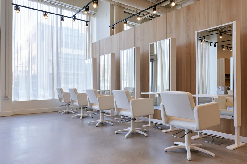 Modern hair salon interior with white styling chairs and mirrors aligned along wooden wall near large windows