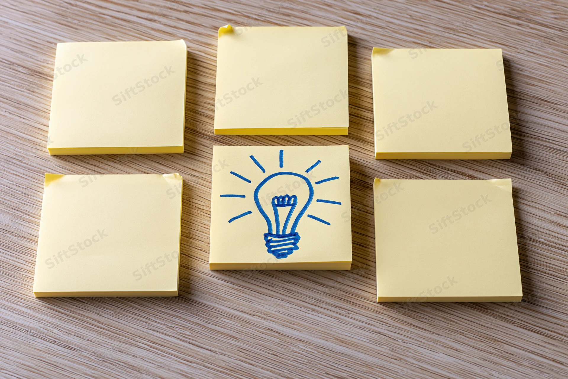 Yellow sticky notes arranged in grid on wooden desk with one featuring blue lightbulb drawing