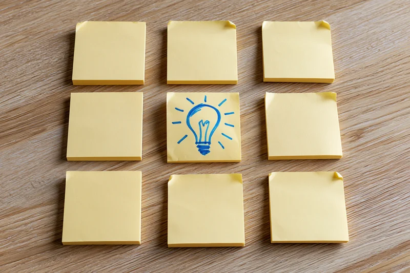 Nine yellow sticky notes in a 3x3 grid on wooden surface with one featuring a blue lightbulb drawing