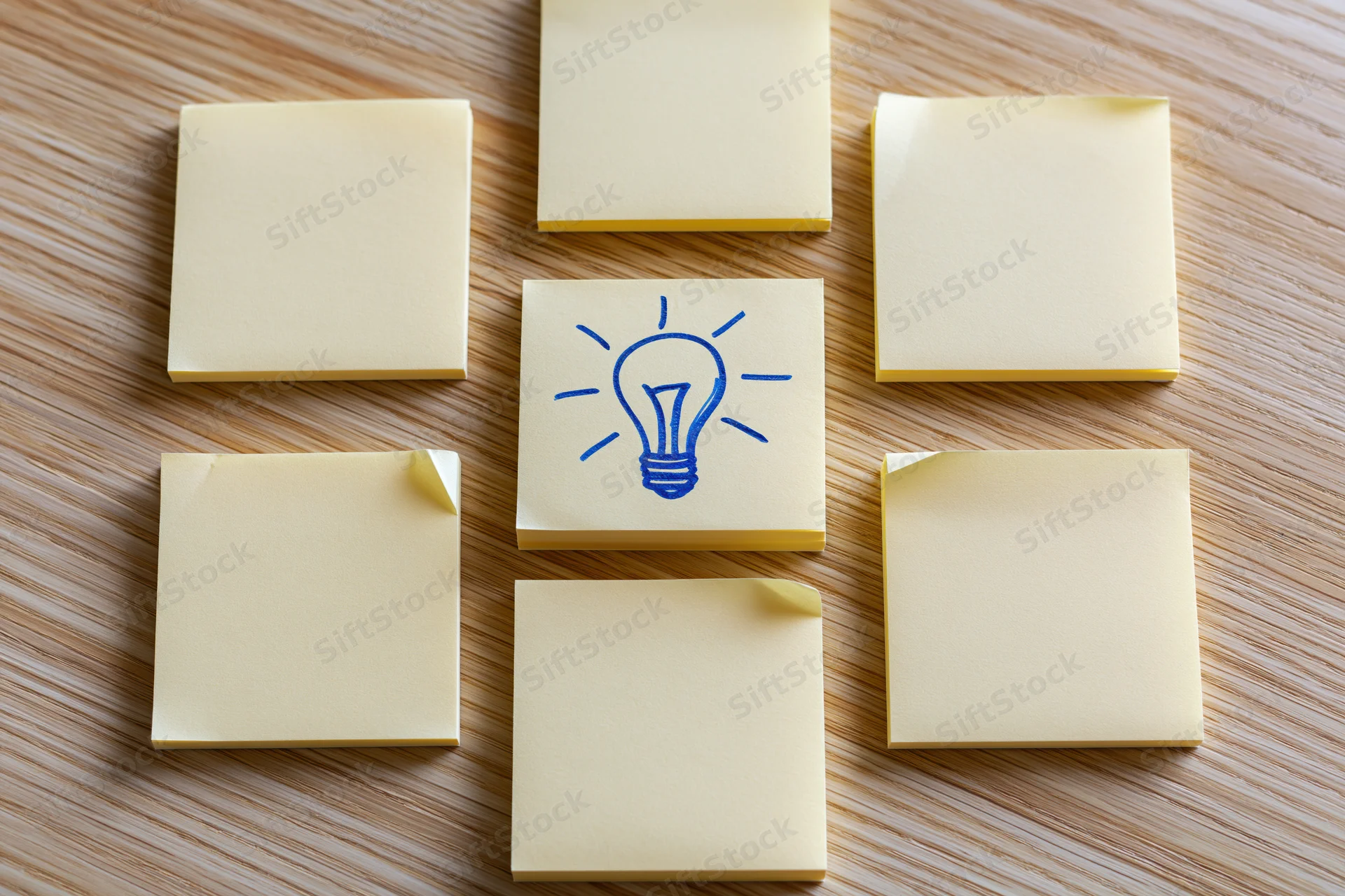Nine yellow sticky notes arranged in a 3x3 grid on a wooden desk with one featuring a blue lightbulb drawing