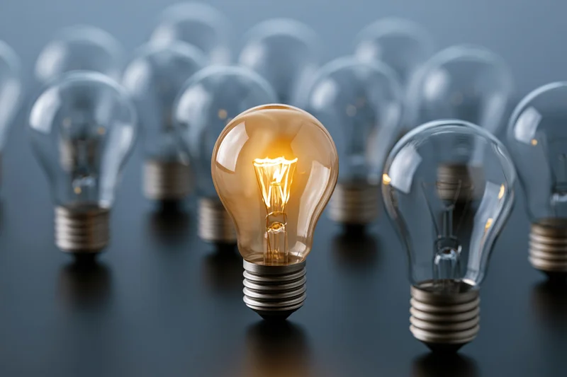 One glowing incandescent light bulb in focus among unlit bulbs on a dark surface
