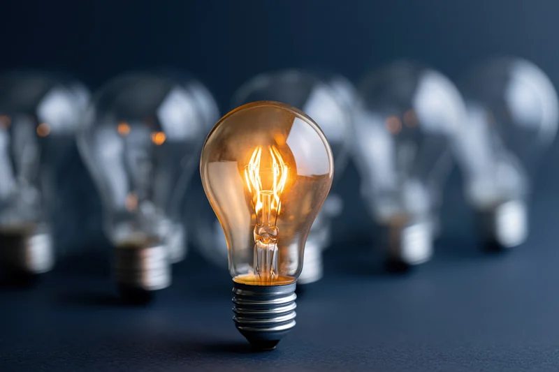 Glowing incandescent lightbulb in focus among unlit bulbs on dark surface