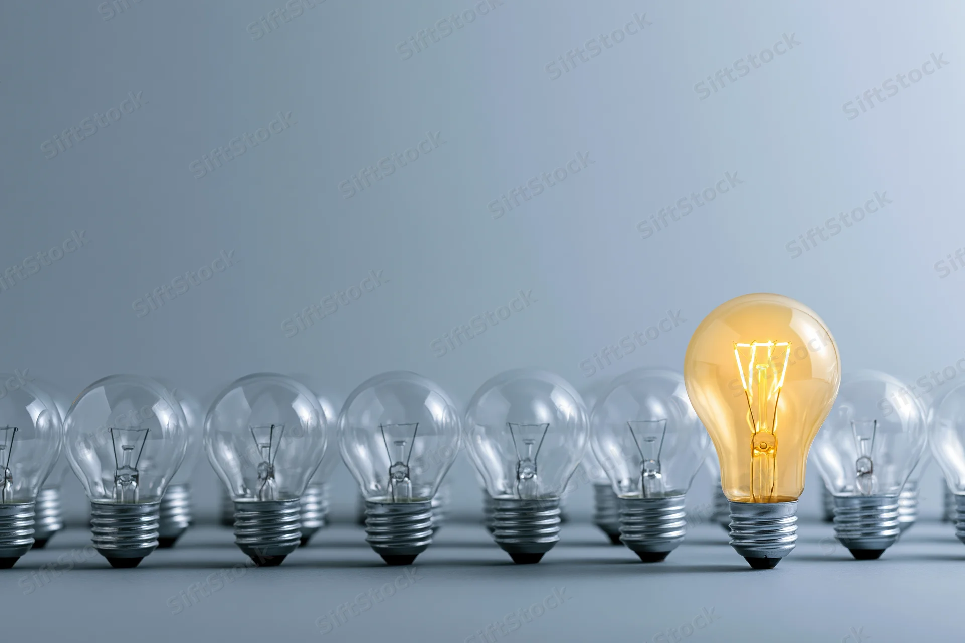One glowing incandescent light bulb standing taller than a row of unlit bulbs on a plain surface