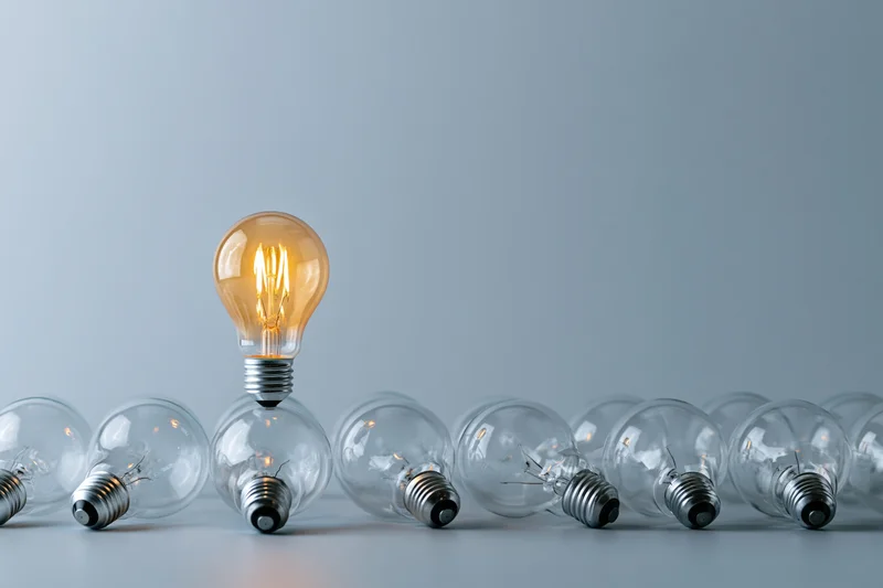 One glowing incandescent light bulb standing upright among unlit bulbs in a row on a gray surface