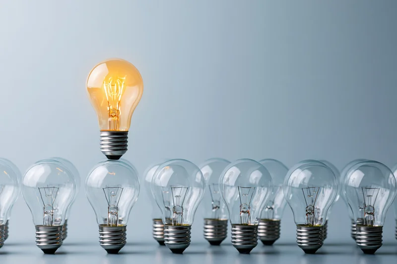 Glowing incandescent lightbulb standing taller than a row of unlit bulbs on a plain surface
