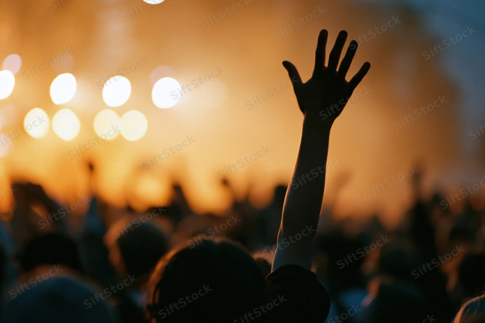 Silhouette of a hand raised high above a blurred crowd at a concert stage with warm glowing lights