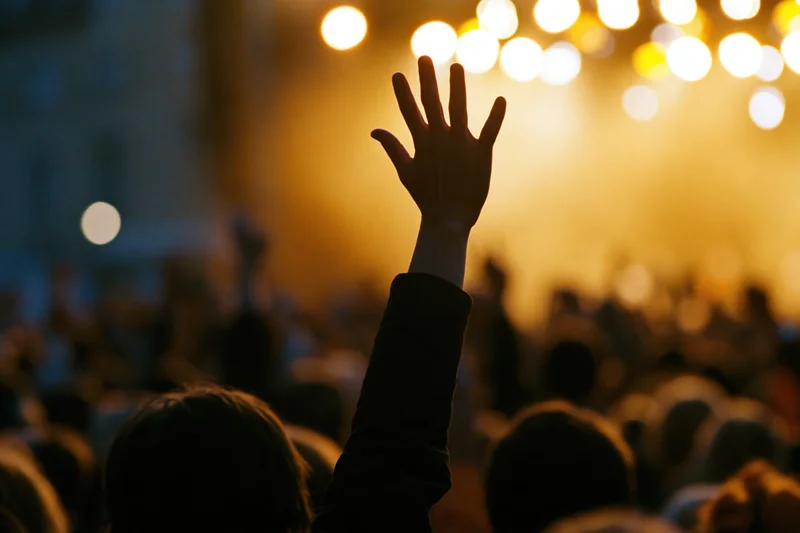 Hand raised in silhouette above a blurred crowd at a concert with stage lights glowing in the background