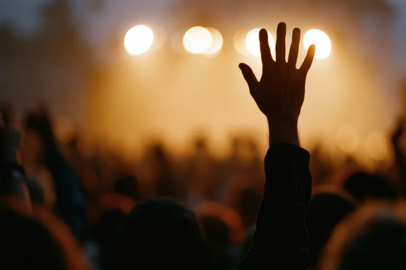 Hand raised in silhouette above a blurred crowd at a concert stage with warm stage lights