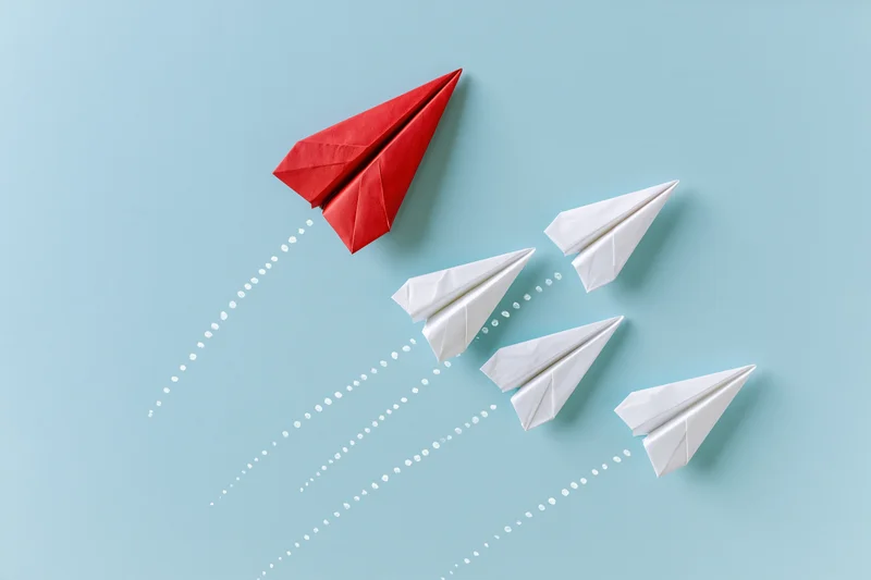 Red paper airplane leading white ones in flight against blue background