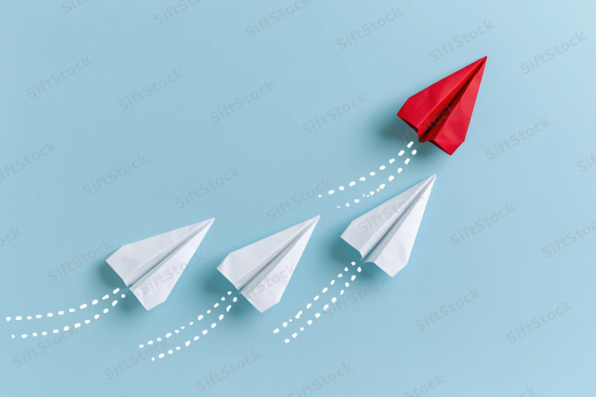 Red paper airplane ascending away from three white ones on a blue background