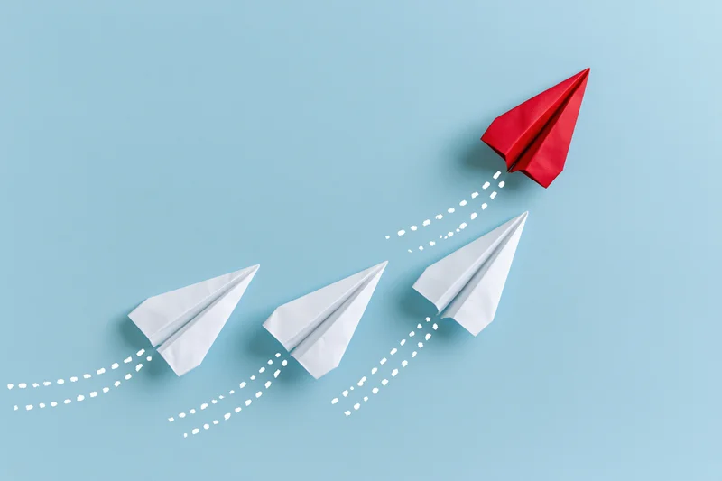 Red paper airplane ascending away from three white ones on a blue background