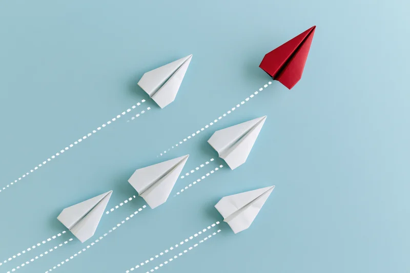 Red paper airplane diverging upward from white ones on blue background