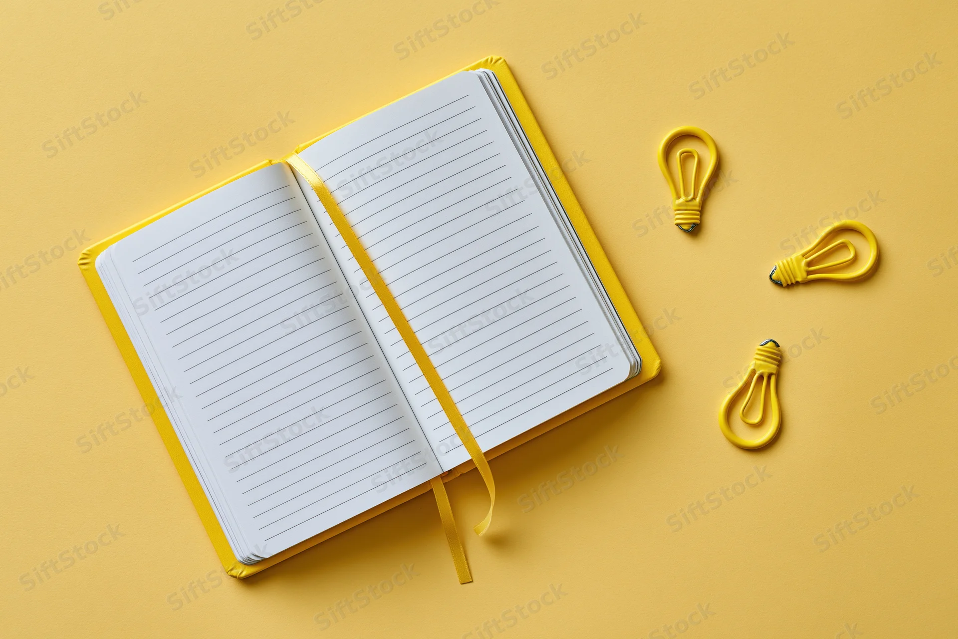 Open blank lined notebook with yellow ribbon and three lightbulb shapes on a bright yellow surface