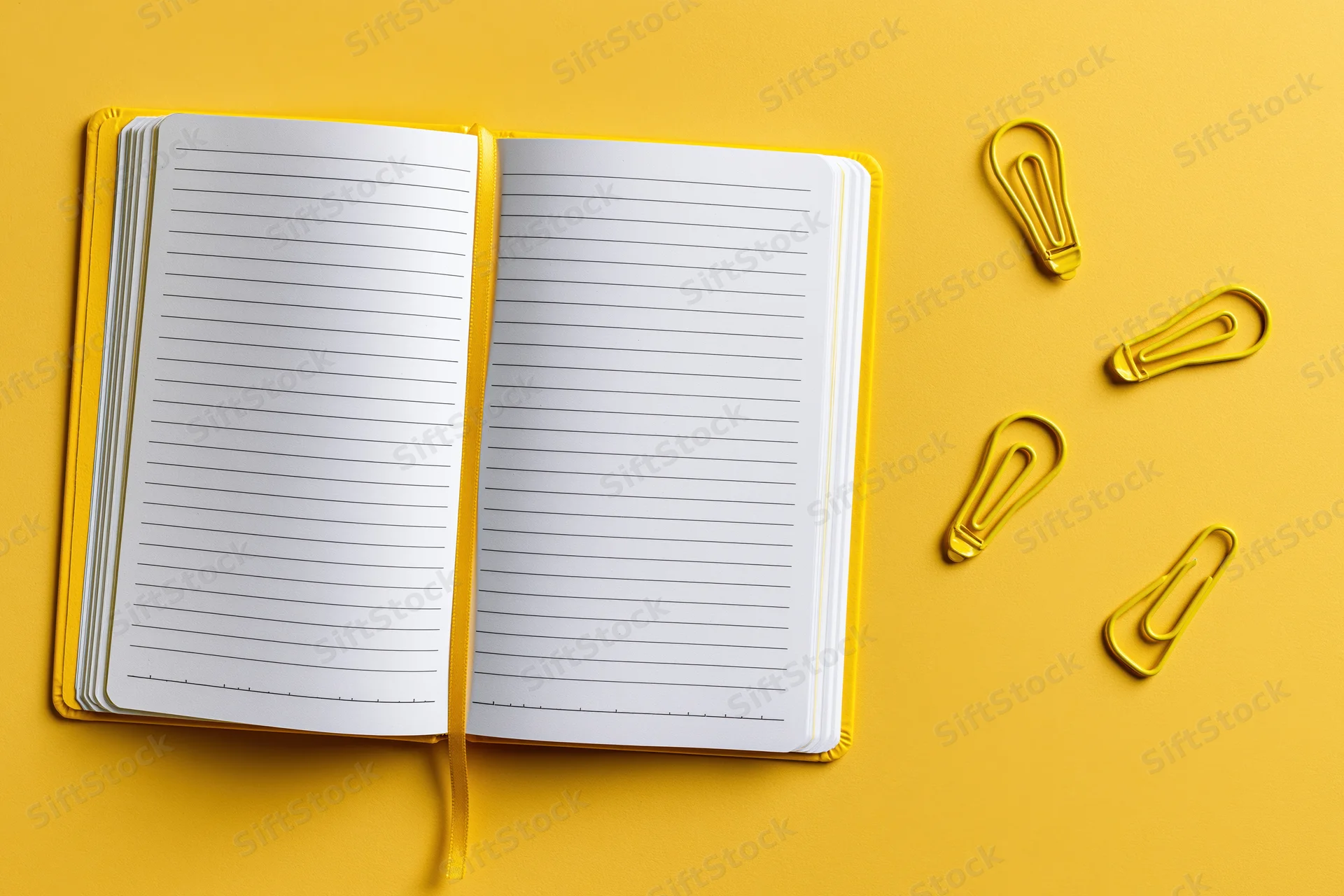 Open blank lined notebook lying flat on bright yellow surface with yellow paperclips shaped like lightbulbs