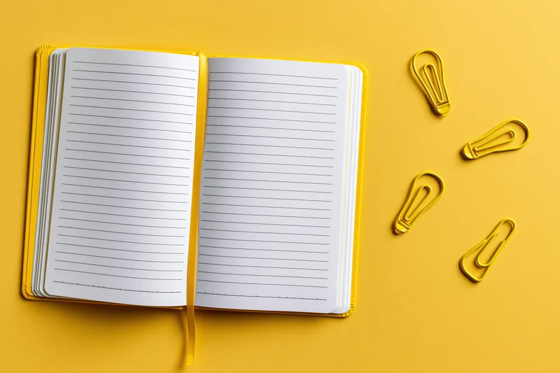 Open blank lined notebook lying flat on bright yellow surface with yellow paperclips shaped like lightbulbs