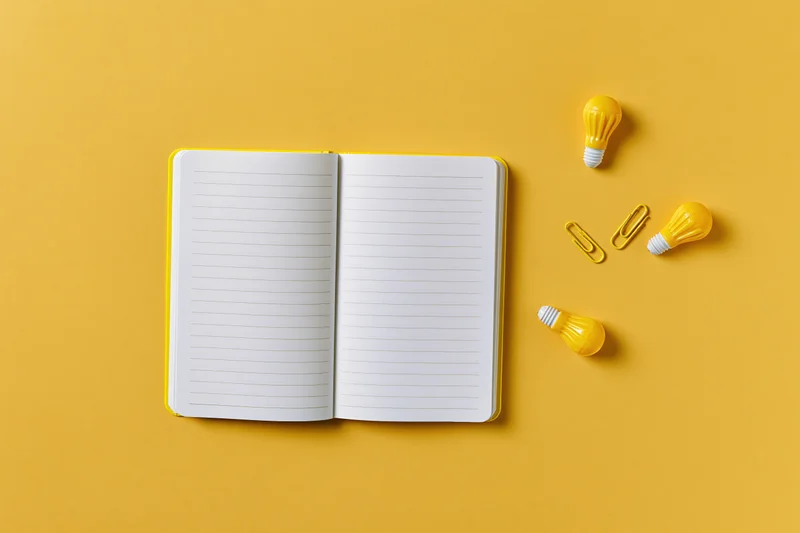 Open blank lined notebook with yellow lightbulbs and paperclips on yellow surface