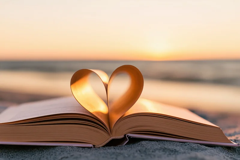 Open hardcover book with pages folded into heart shape on sandy beach at sunset