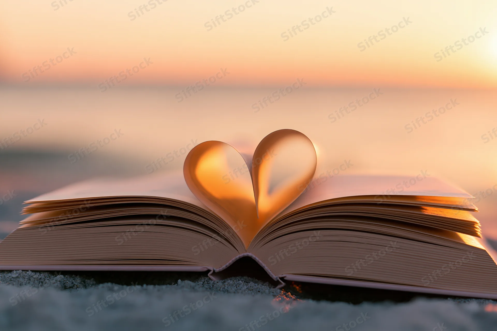 Open book with pages folded into heart shape on sandy beach at sunset