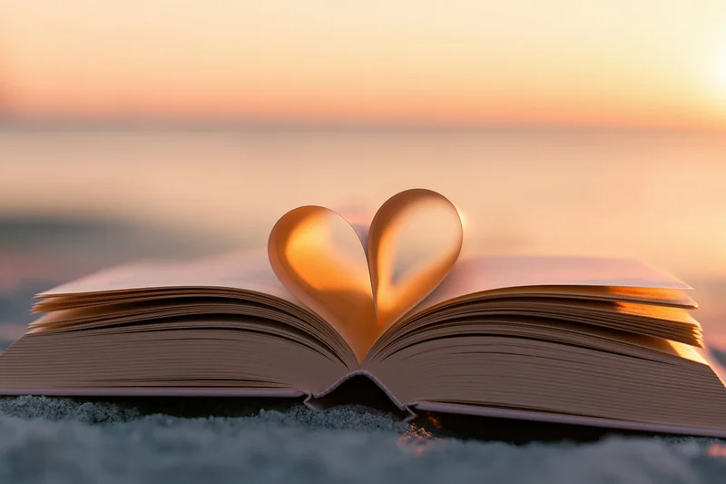 Open book with pages folded into heart shape on sandy beach at sunset