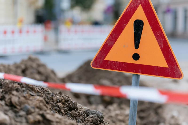 Triangular warning sign with exclamation mark on construction site surrounded by dirt and striped barrier tape