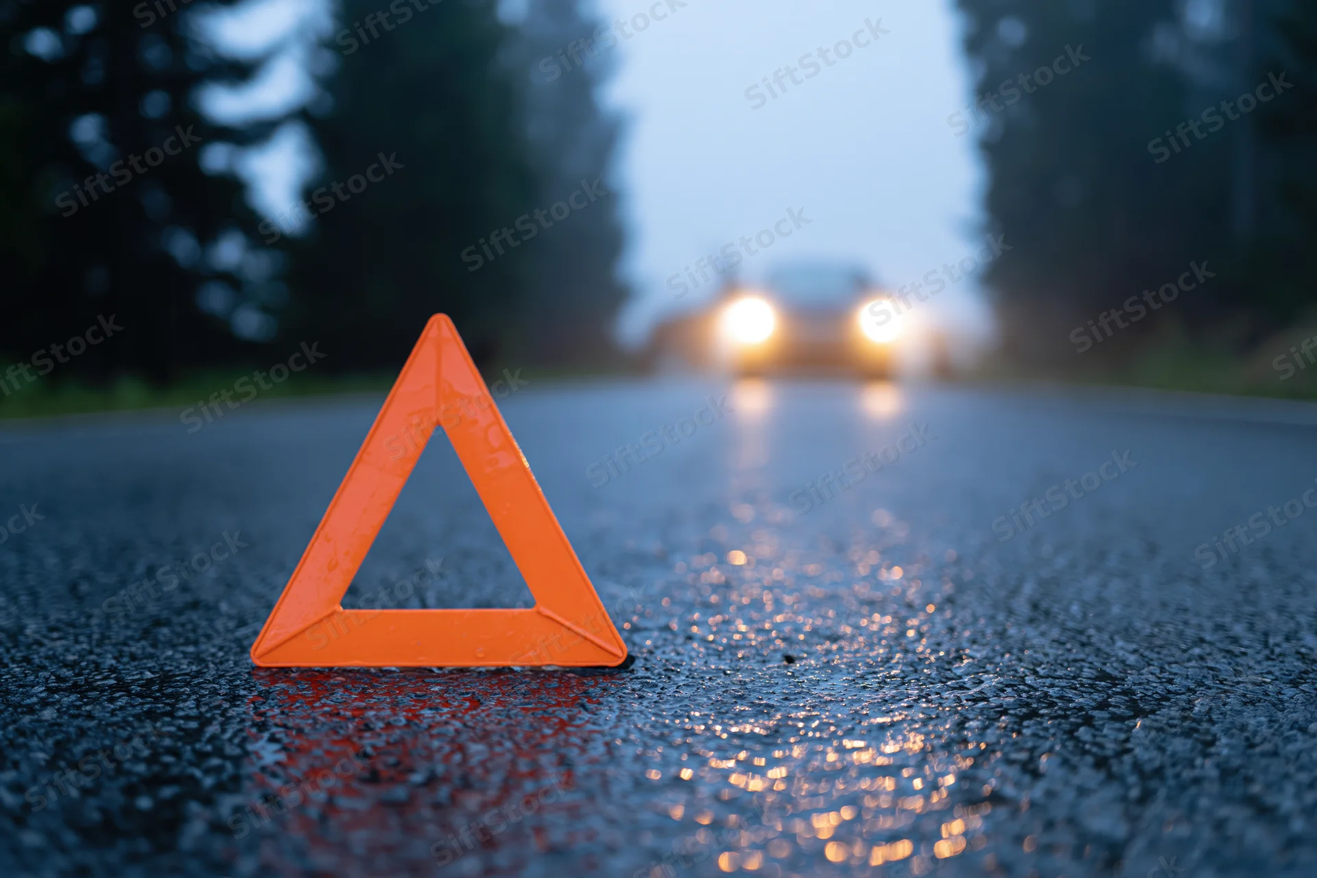 Orange reflective warning triangle on wet asphalt road at dusk with approaching car headlights