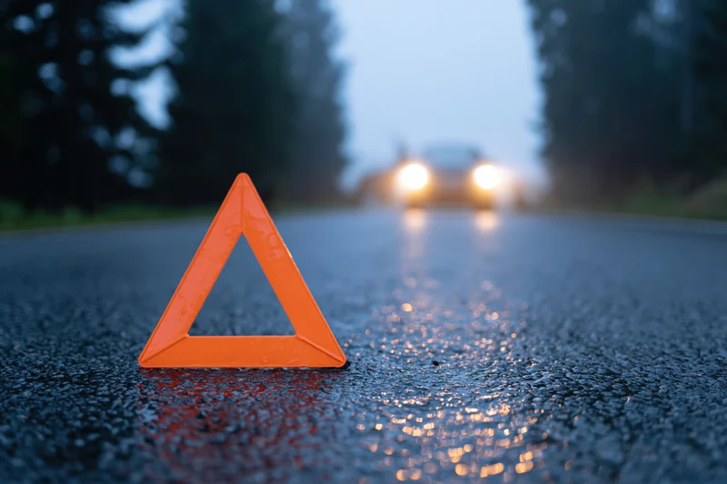 Orange reflective warning triangle on wet asphalt road at dusk with approaching car headlights