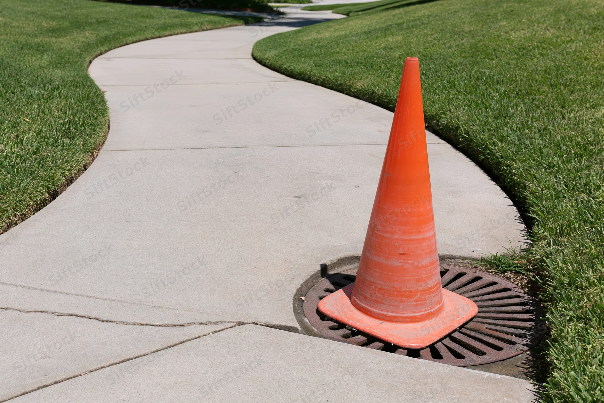 Orange traffic cone placed on a storm drain grate beside a curved concrete sidewalk in a grassy park area