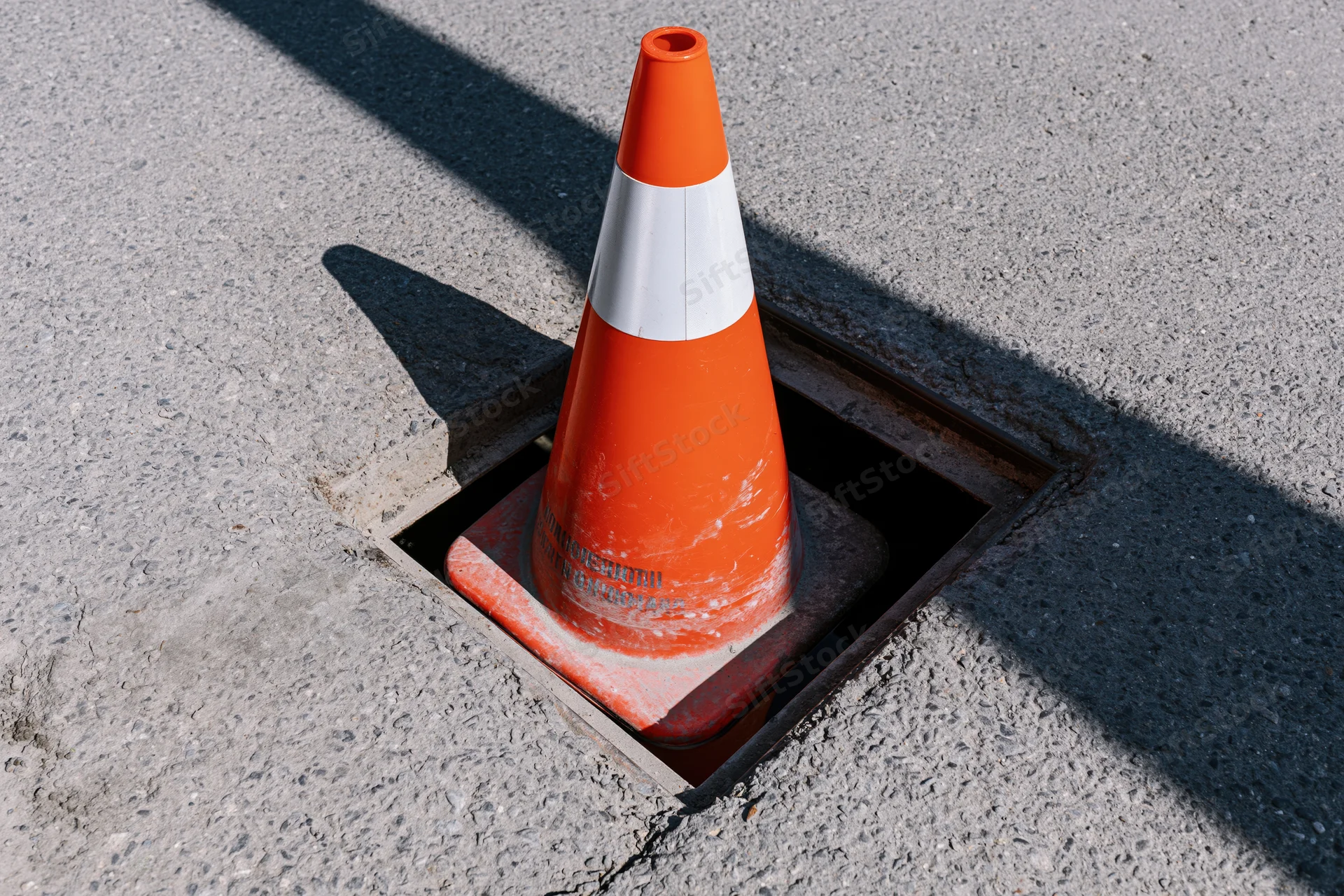 Orange traffic cone with reflective white band sitting inside a square drain cover on asphalt pavement