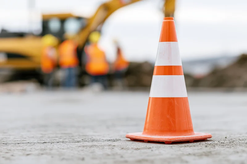 Orange traffic cone with white reflective bands on pavement at construction site with workers and excavator in background