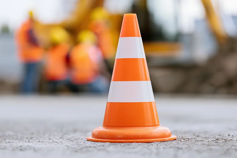 Orange traffic cone with white reflective bands on asphalt at construction site with workers and machinery in background