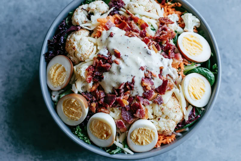 Overhead close-up of a large salad bowl filled with roasted cauliflower, bacon, hard-boiled eggs, shredded cheese, carrots, and leafy greens, topped with creamy dressing