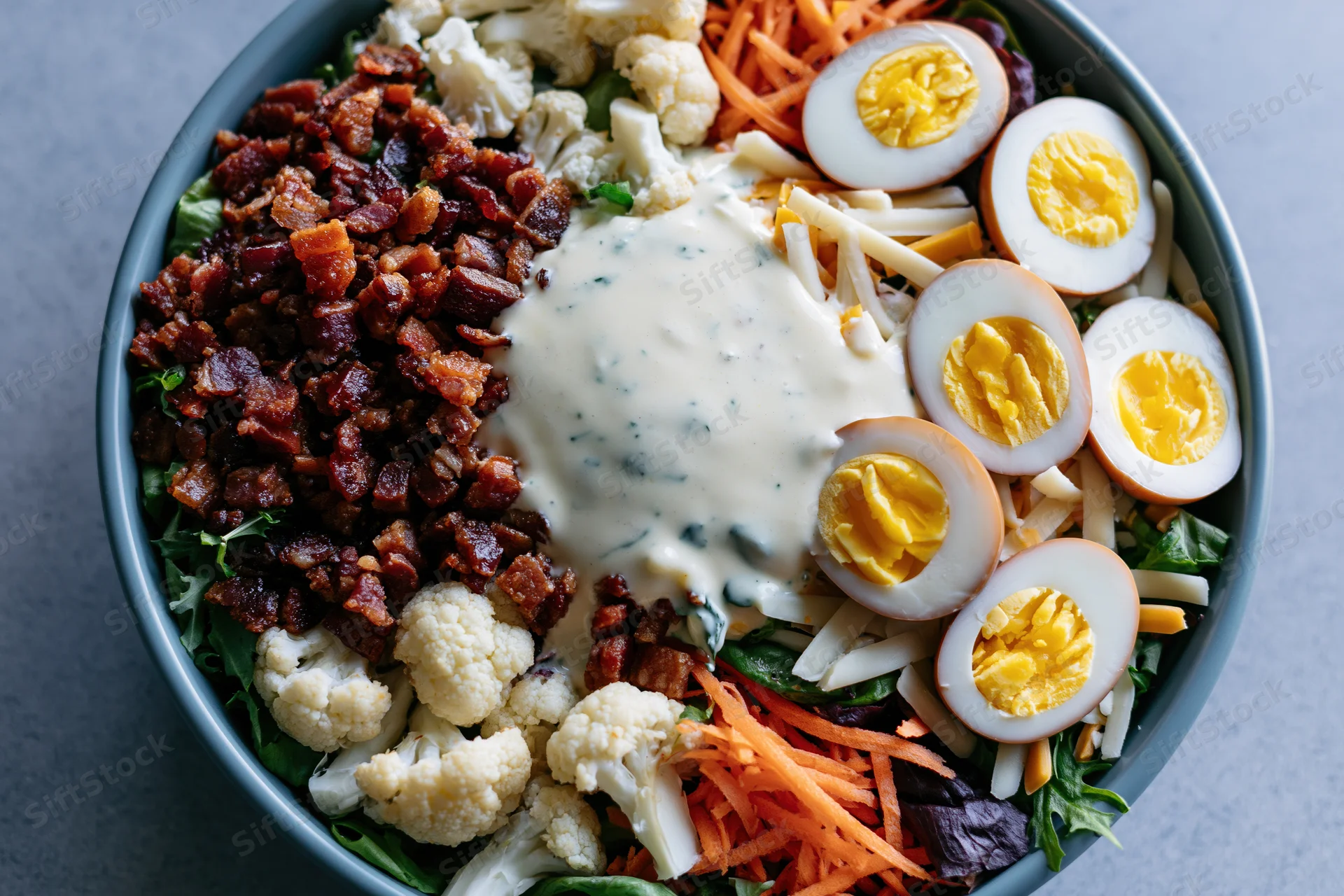 Overhead close-up of a large salad bowl filled with bacon, hardboiled eggs, shredded cheese, cauliflower, carrots, lettuce and blue cheese dressing