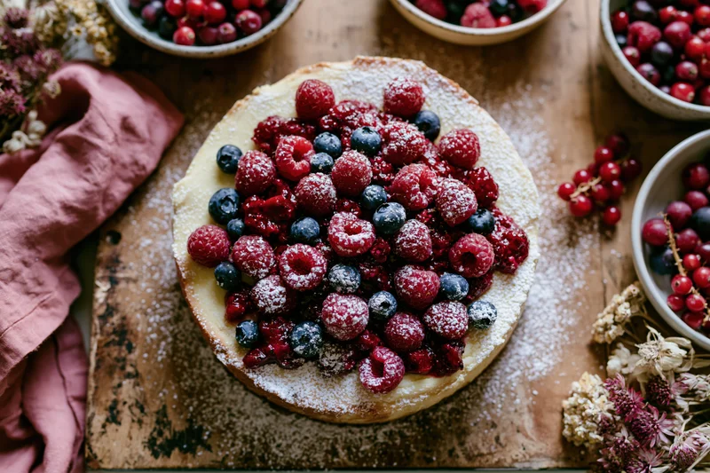 Round cheesecake topped with fresh berries and powdered sugar on wooden board with bowls and dried flowers
