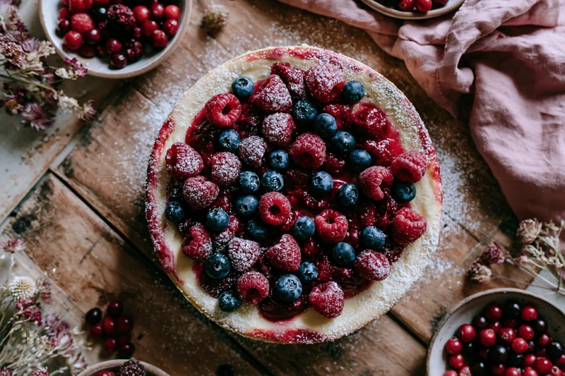 Round cheesecake topped with fresh raspberries and blueberries on a wooden table with bowls of berries and dried flowers around