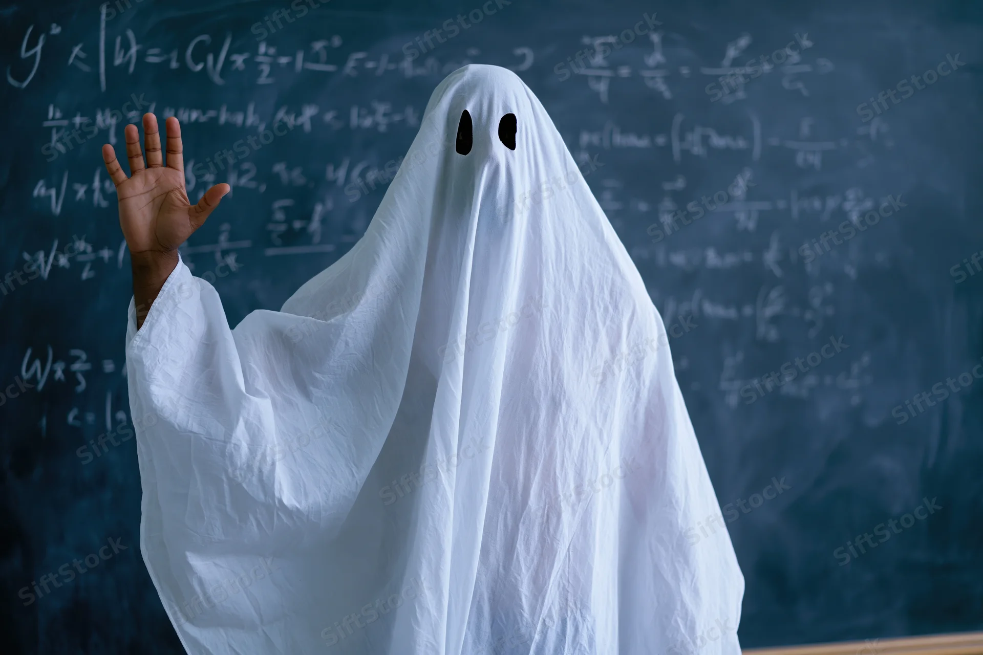 Ghost in white sheet costume raising hand in front of blackboard filled with math equations