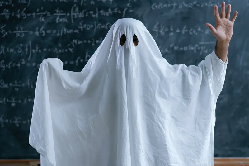Student in white sheet ghost costume raising hand in front of chalkboard filled with math equations