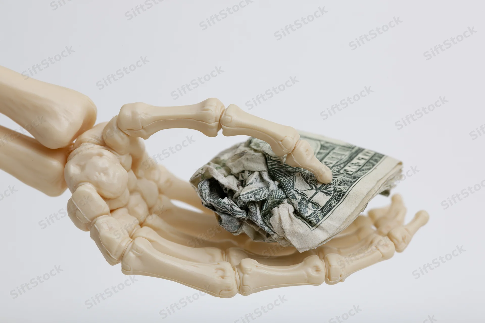 Skeleton hand gripping a crumpled stack of dollar bills against a white background