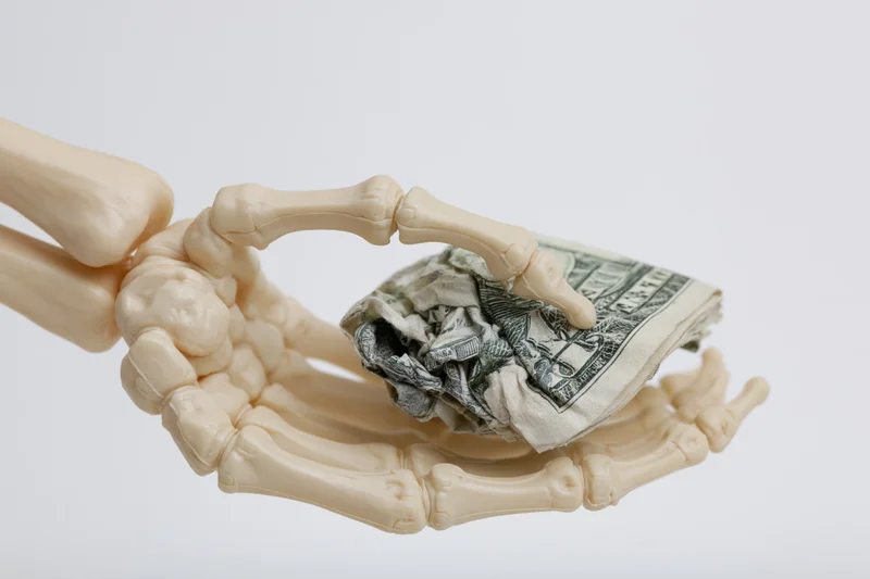 Skeleton hand gripping a crumpled stack of dollar bills against a white background