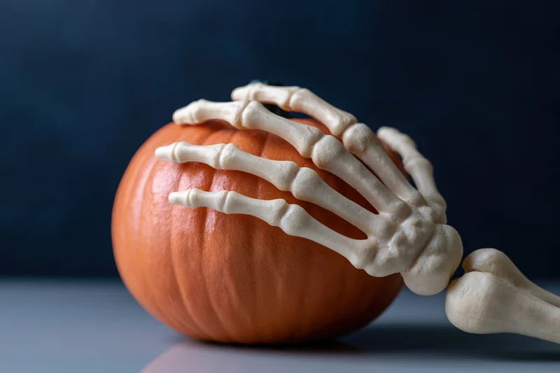 Plastic skeleton hand gripping an orange pumpkin on a dark blue surface