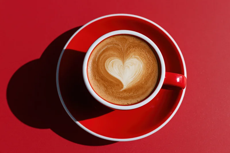 Red and white ceramic cappuccino cup with heart-shaped latte art on matching saucer against red background