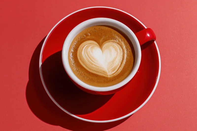 Red and white ceramic cappuccino cup with heart-shaped latte art on matching saucer against red background